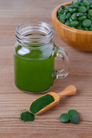 sun chlorella tablets in wooden bowl, sun chlorella powder form in scoop in front of glass jar style mug full of water with sun chlorella mixed in