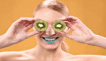 a head and shoulder picture of a blonde lady holding pieces of kiwi fruit slices in front of her eyes and wearing blue lipstick