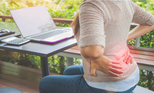 image of a female holding her lower back though in pain, while sitting in front of her laptop computer