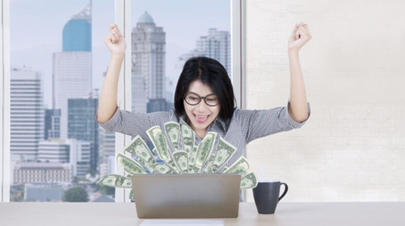 excited lady has money popping up from her desktop computer in front of her